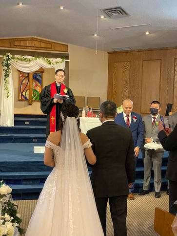 A bride being led down the aisle by her father with the wedding officiant on stage and the groom + best man on the right.