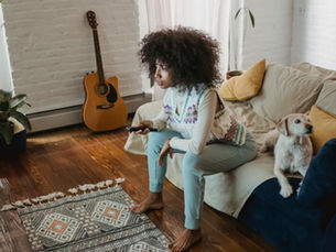 Woman sits on a couch with a remote, a dog beside her. A guitar and plant in background. Cozy room with colourful rug and bright light.