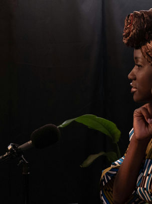 A person with a patterned headscarf adjusts earrings near a microphone. They're in a dimly lit room with a plant in the background.