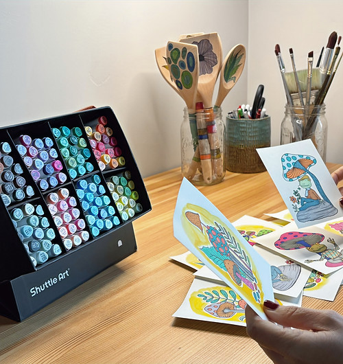 A case brimming with a wide assortment of colorful art markers sits on a wooden desk, surrounding a collection of whimsical, hand-drawn illustrations, primarily featuring mushrooms and frogs. A person's hands are holding up two of these vibrant artworks, showcasing their artistic creations.