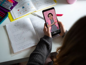 Person in gray sweater holding a smartphone showing a video. Open notebooks and colorful pens on desk. Bright, casual setting.