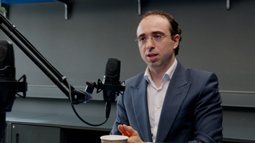 Loran wearing a dark jacket while speaking near a studio microphone on set.