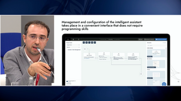 Loran Jacobs sits at a panel next to a slide detailing no-code management of intelligent assistants.