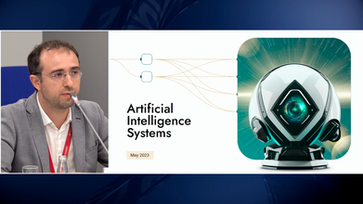 Loran Jacobs is seated at a table, holding a microphone next to a slide titled "artificial intelligence systems".