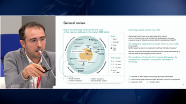 Chest-up view of Loran Jacobs addressing the forum beside a projection slide titled "General review".