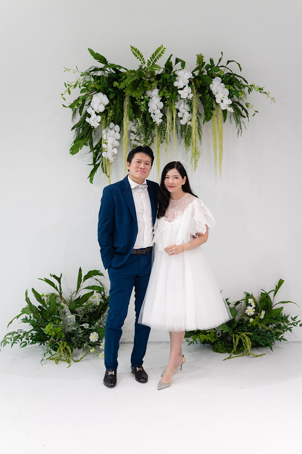 Bride in white dress and groom in blue suit pose together before a lush green-and-white floral backdrop, smiling softly.