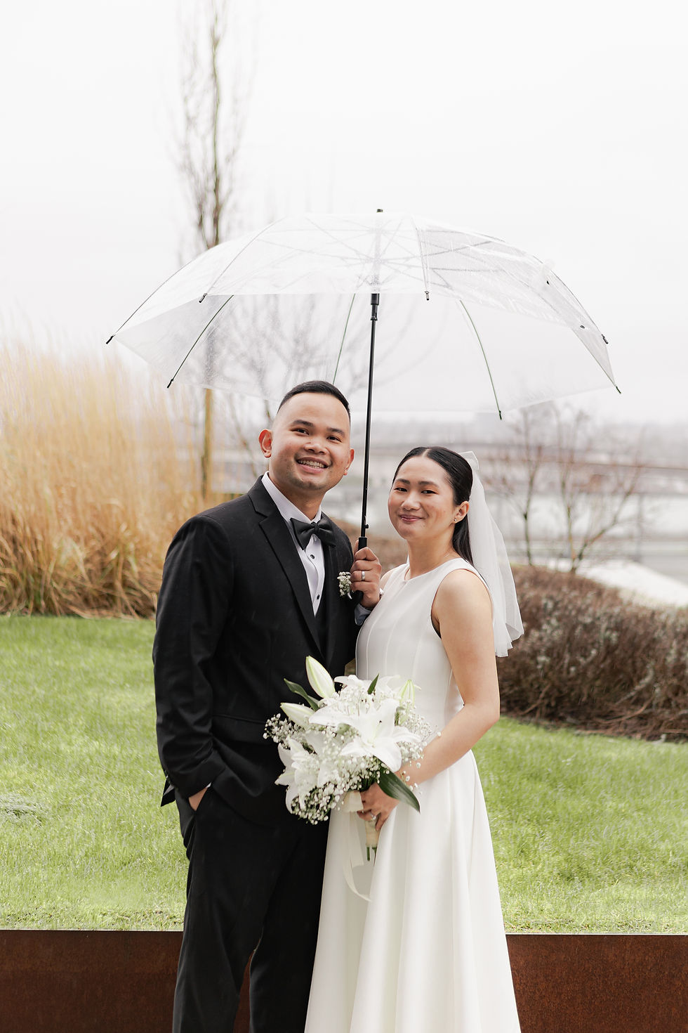 Smiling bride and groom in wedding attire stand outdoors under a clear umbrella, holding a white bouquet.