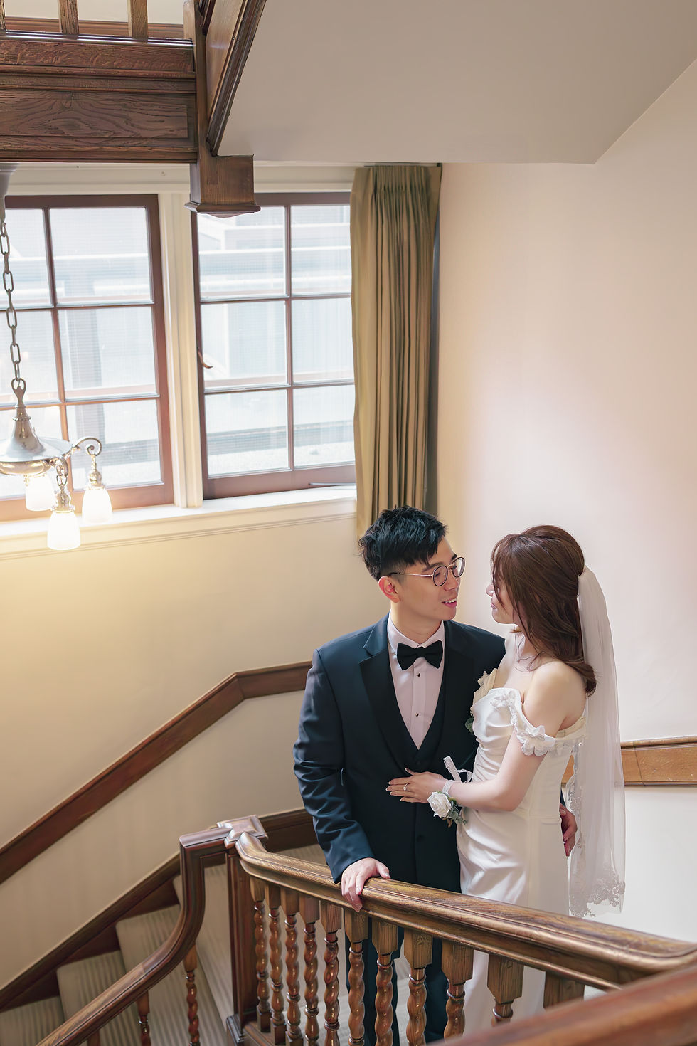 Bride and groom in wedding attire gaze at each other on a wooden staircase in a bright, elegant home.