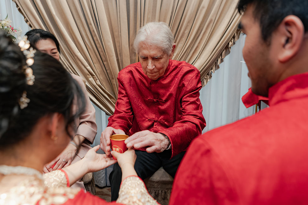 Elderly man in a red silk jacket receives a red cup from family during a solemn indoor ceremony with beige drapes.