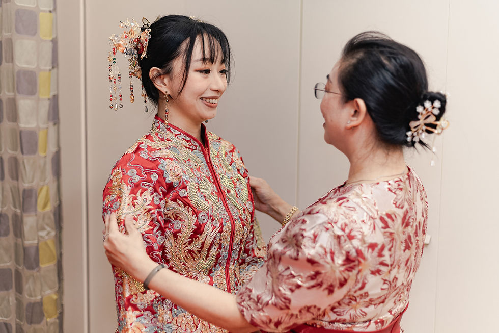 Smiling woman in ornate red Chinese dress and jeweled hairpiece is being adjusted by an older woman indoors.