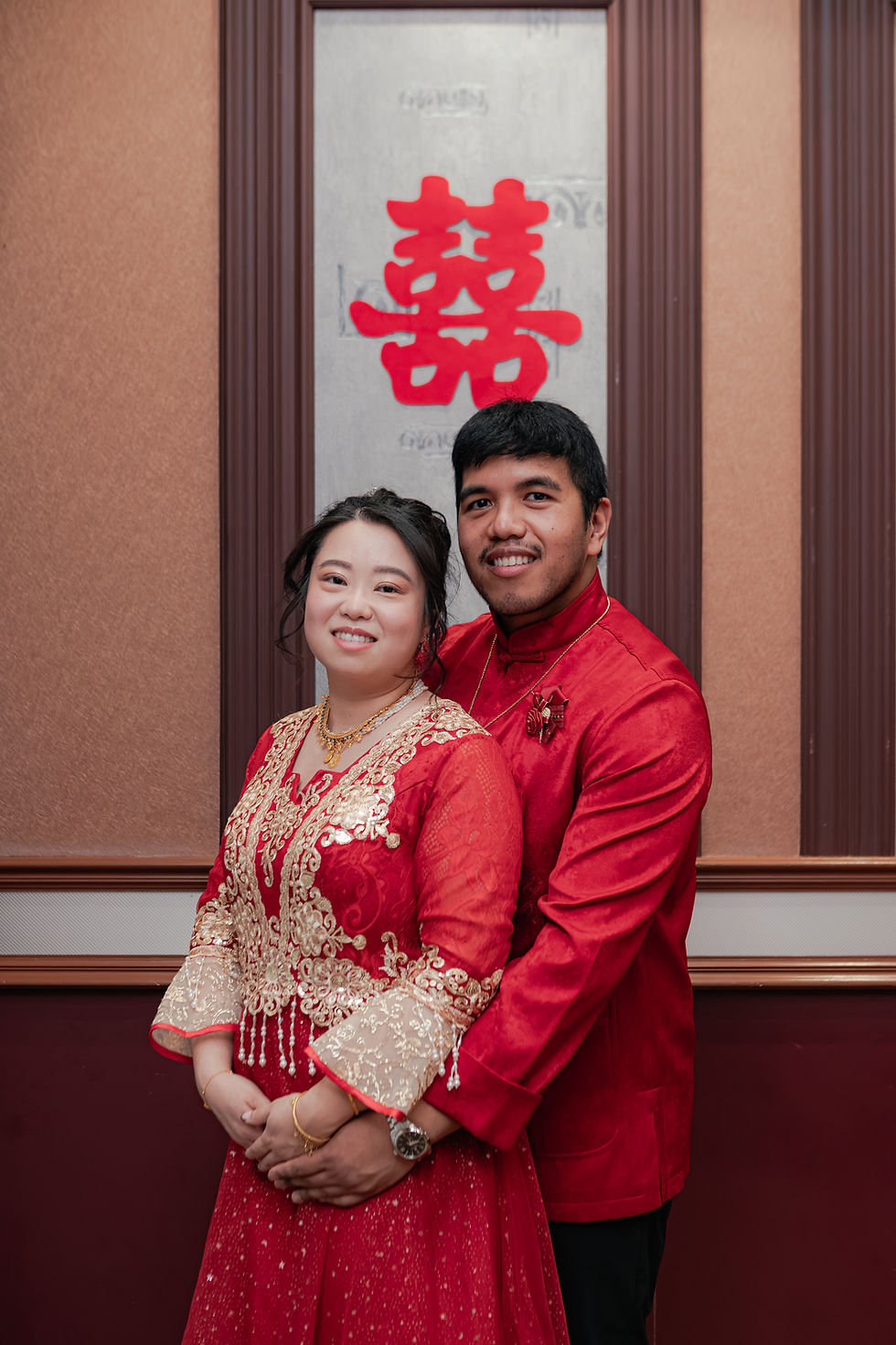 Smiling couple in red traditional wedding attire pose before a red double-happiness sign on a paneled indoor wall.