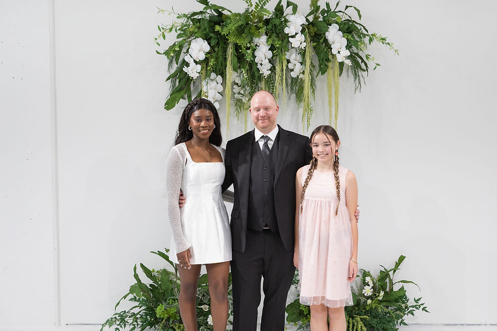 Three people pose smiling under a white-and-green floral arch, dressed in formal black, white, and pink outfits.