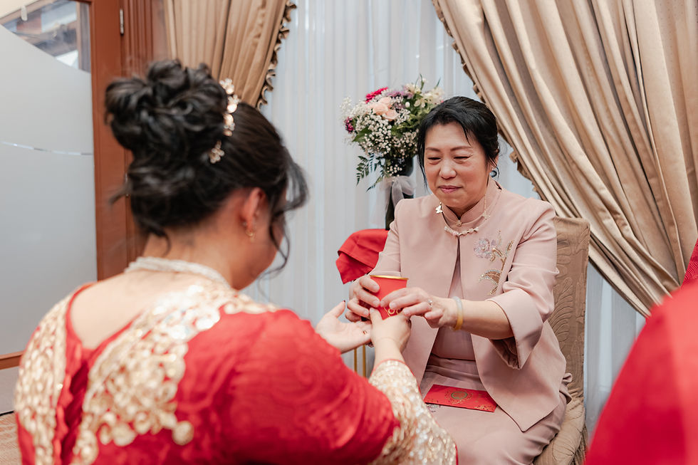 Woman in pink hands a red envelope to a woman in a red embroidered dress in a elegant curtained room with flowers.