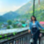 A smiling woman sitting on a balcony railing in front of a mountain town with colorful rooftops and dense green forested hill