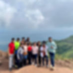 Eleven people standing on a mountain ridge, smiling at the camera, with green hills and cloudy sky in the background.