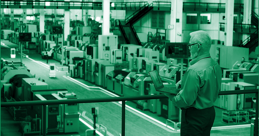 man in business attire standing on an upper deck looking down at a manufacturing facility holding a laptop computer