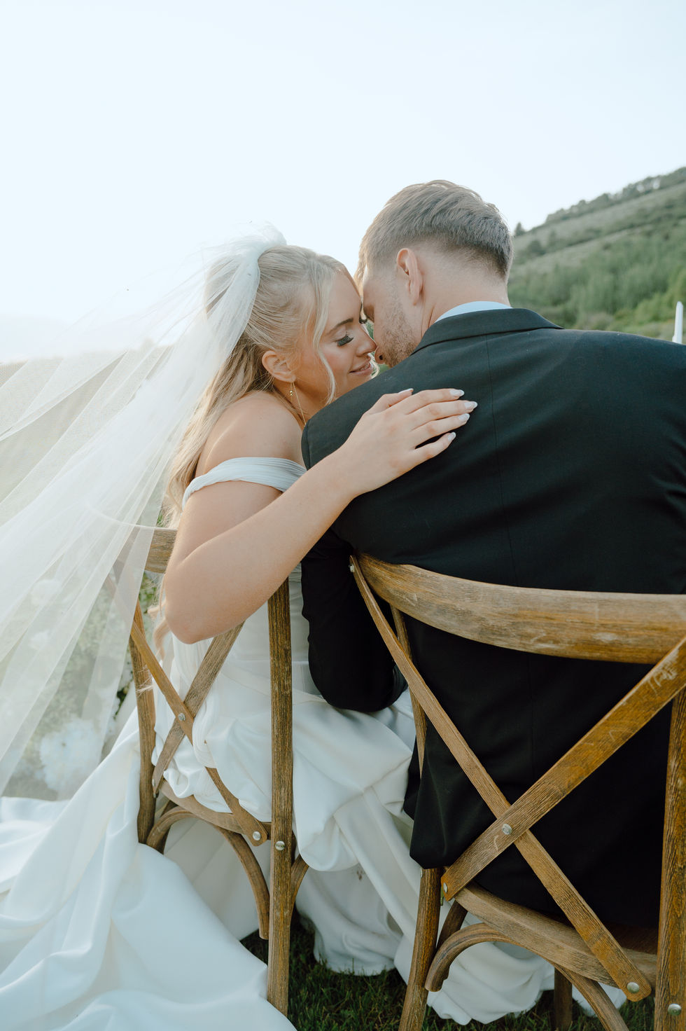 Bride and groom sitting closely on wooden chairs.