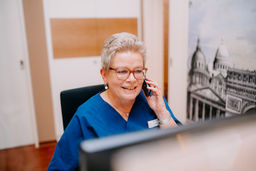 Lächelnde Frau in blauer Arbeitskleidung telefoniert am Schreibtisch.