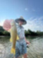 Person in a floral hat and glasses holding a fish, standing in a river. They appear joyful. Blue sky and trees in the background.