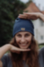 Portrait von braunhaariger Frau die frontal in die Kamera schaut. Sie lächelt und rahmt ihren Kopf mit beiden Händen, dabei trägt sie die stoff.werk Baumwollmütze in dunkelblau.