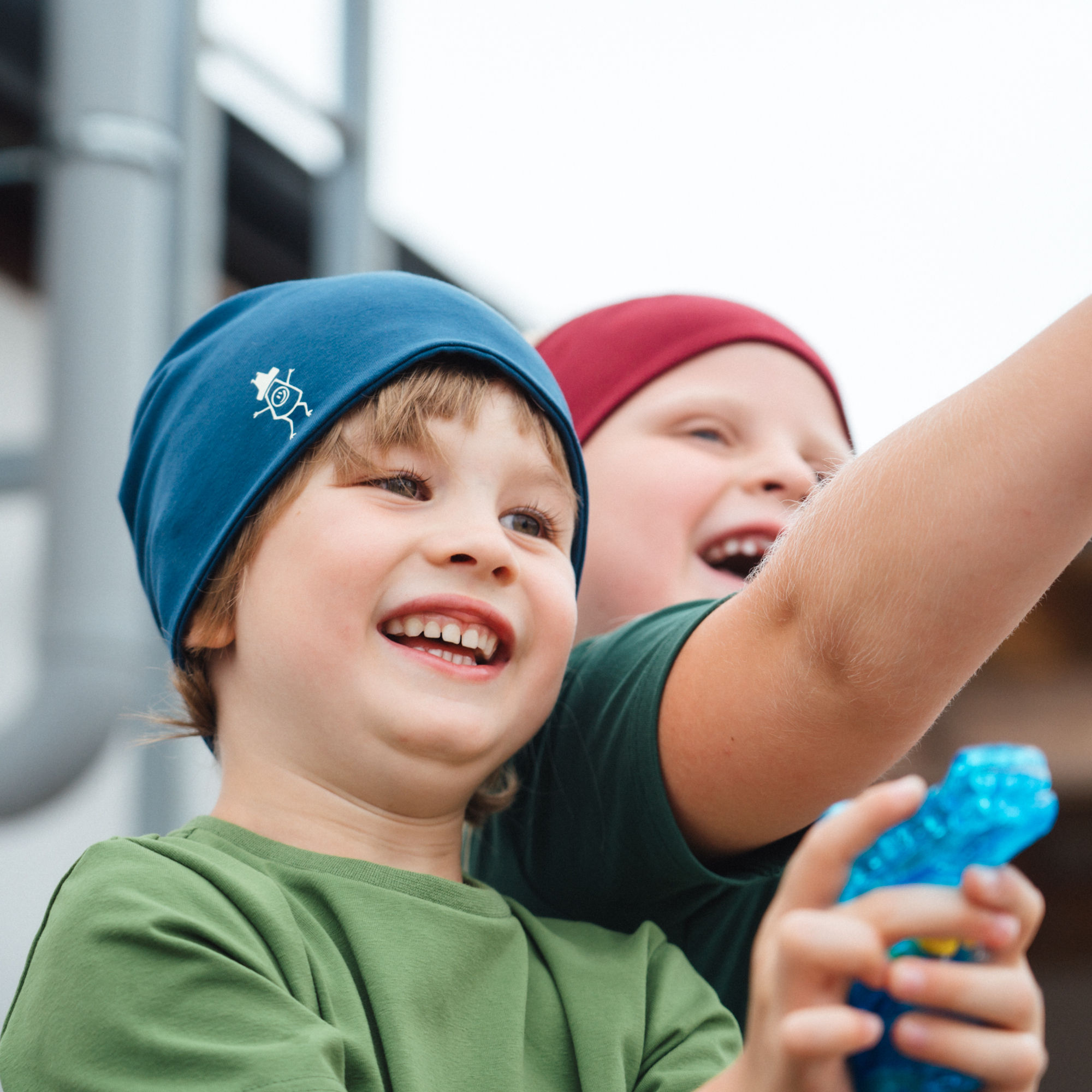 Ein Mädchen und ein Bub spielen mit Wasserpistolen und lachen fröhlich - der Bub trägt eine blaue Baumwollhaube zu einem Hellgrünen T-Shirt und das Mädchen eine rote Haube zu einem dunkelgrünen T-Shirt.-