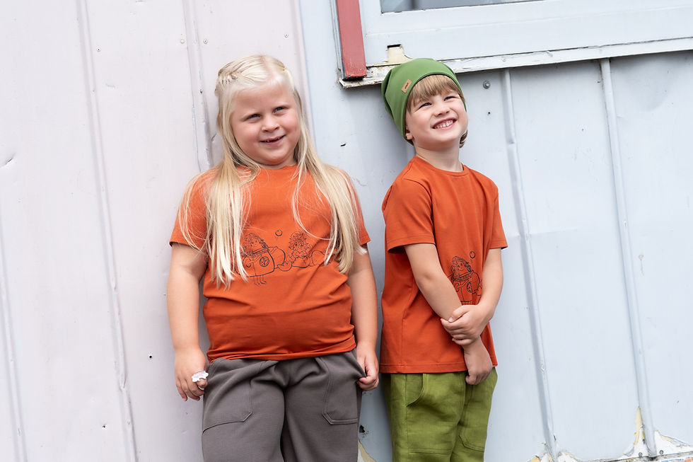 Blondes Mädchen und braunhaariger Bub tragen beide ein rotes T-Shirt und lehnen gegen eine weiße Wand.