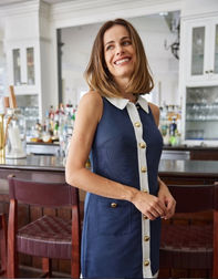 Gabriela Dias in a sleeveless navy dress with white trim and gold buttons, standing at a bar with wooden chairs and glass shelves. • Gabriela Dias com vestido azul-marinho sem mangas, com detalhes brancos e botões dourados, em frente a um bar com cadeiras de madeira e prateleiras de vidro.