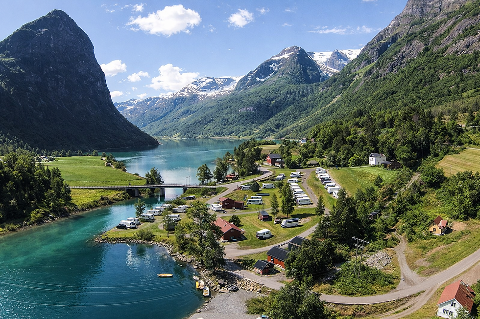 Mountainous landscape with a turquoise lake, caravans, and red cabins in a green valley. Snow-capped peaks under a blue sky. Peaceful mood.