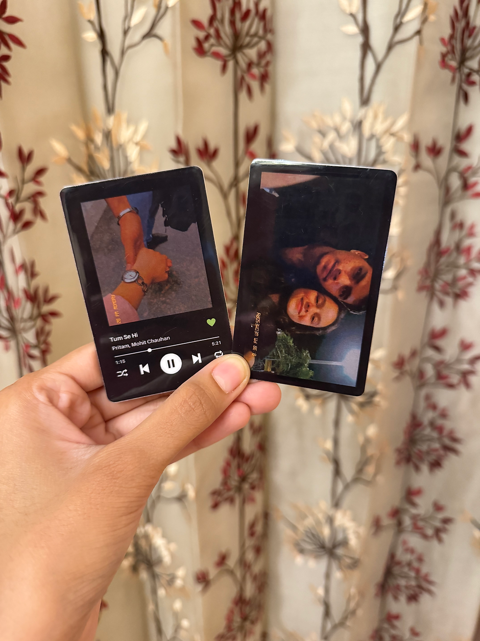 A hand holds two black, rectangular personalized art gifts by Ecstatic Art, likely Wallet Card Art. One card features a photo of a couple holding hands, overlaid with a custom Spotify song code for "Tum Se Hi." The other card shows a photo of a couple smiling. These are examples of unique art gifts and affordable art India.