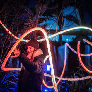 A woman in a winter hat and jacket interacts with a large, illuminated neon light sculpture in a dark, outdoor setting; the sculpture forms abstract shapes, curves, and lines in shades of orange - designed by Light Artist Frankie Boyle
