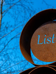 Escultura metálica ao ar livre com a palavra “Listen”, recortada sobre fundo azul e árvores desfocadas.