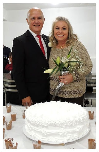 Casal sorrindo perto de bolo branco e arranjo floral, em evento social. My Fraternity News