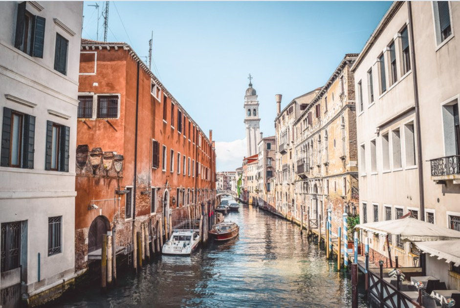 Canale veneziano con edifici storici, barche ormeggiate e campanile sullo sfondo in una giornata luminosa.