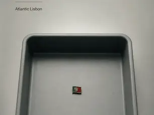 Capa editorial minimalista com um tabuleiro de segurança cinzento e uma pequena bandeira portuguesa ao centro, sob luz fria.