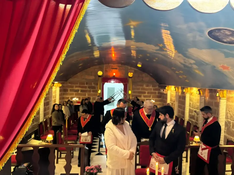 Momento cerimonial numa loja maçónica com cortina vermelha, teto estrelado, velas acesas e participantes reunidos em torno do casal.