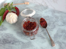 A jar of homemade chilli jam with a spoon beside it, served with fresh red chillies and red peppers.