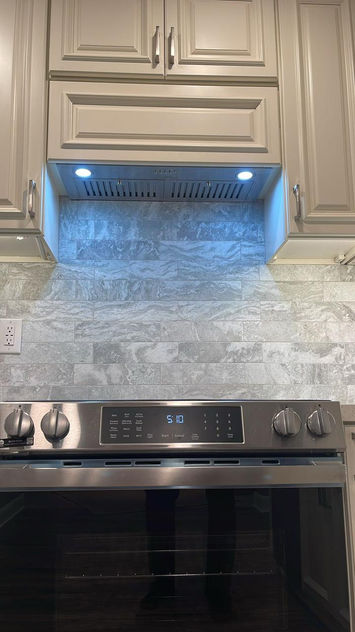 Complete kitchen renovation featuring a transformation from dark wood cabinets and granite countertops to a bright, modern design with cream shaker-style cabinets, quartz countertops, stainless steel appliances, gray stone backsplash, and wood-look flooring
