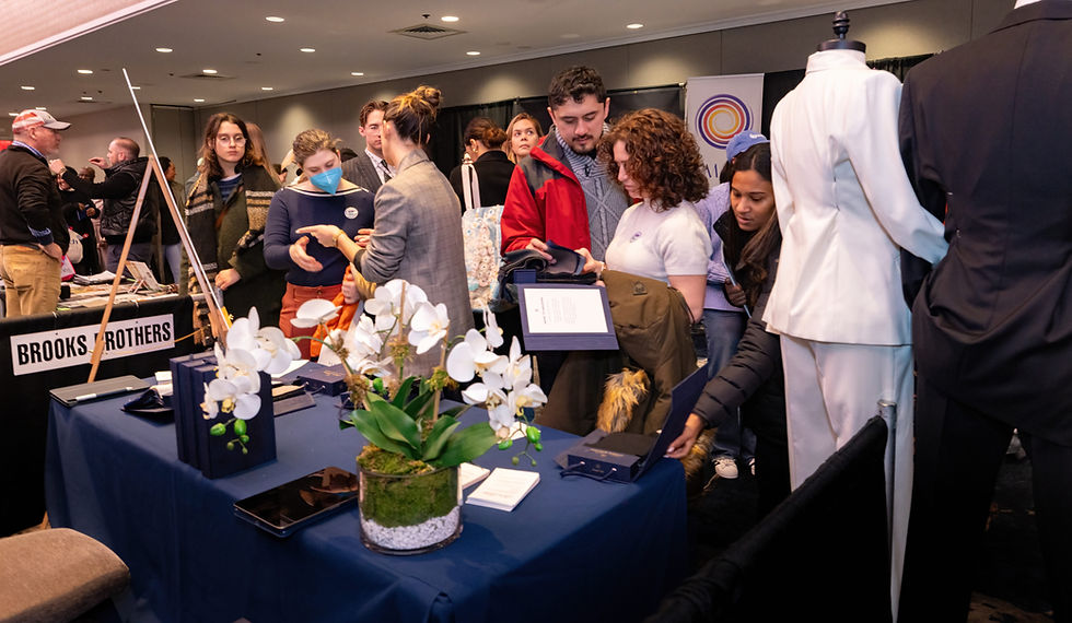 Attendees stand around a table at a wedding show, featuring a man in a suit discussing exhibitor offerings.