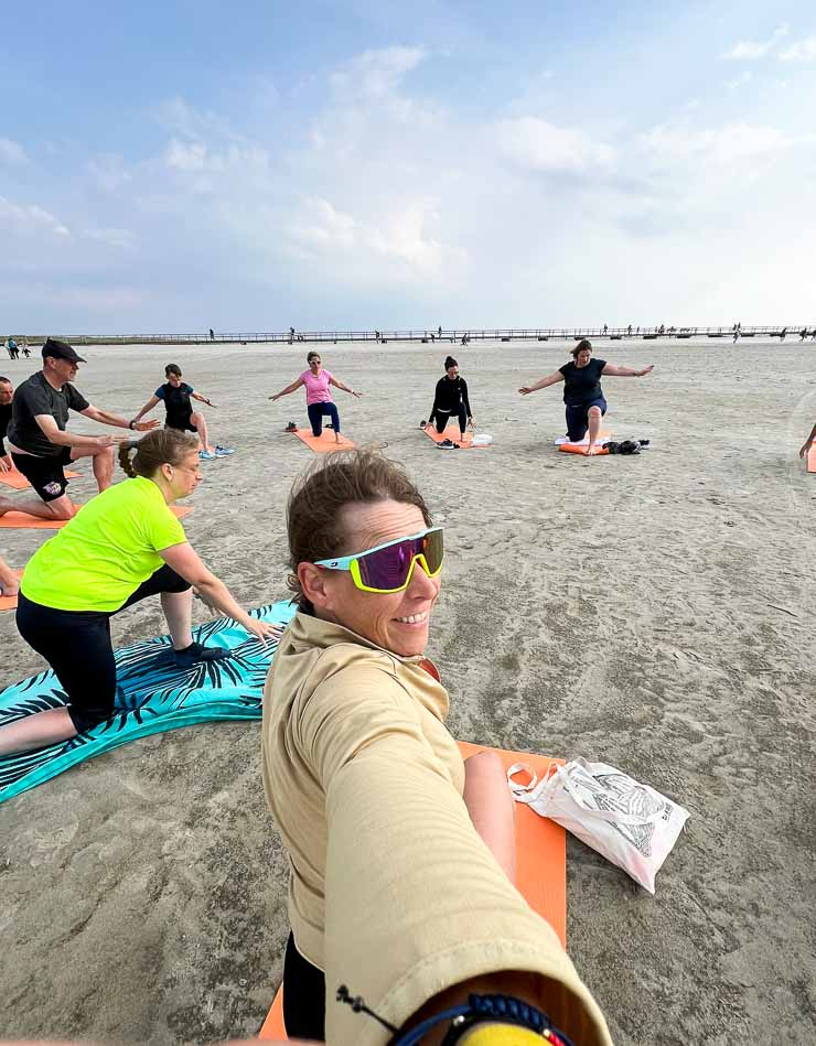 Gruppe von Menschen beim Yoga am Strand auf Matten. Person im Vordergrund macht Selfie und trägt bunte Sonnenbrille. Himmel bewölkt.
