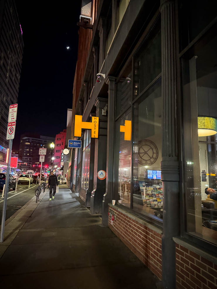 Nächtliche Straßenszene mit einer Person auf dem Gehweg. Gelbes "Hostel"-Schild an einem Gebäude. Rote Lichter der Autos im Hintergrund.