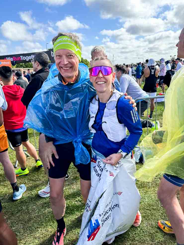 Zwei Personen bei einem Marathon, in bunten Outfits mit blauen Regenponchos und Sonnenbrillen. Im Hintergrund viele Teilnehmer und ein Schild "We Run Together".