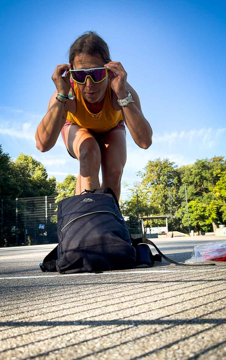 Person mit gelbem Top und Sonnenbrille beugt sich über schwarzen Rucksack im Park. Sonniger Tag, blauer Himmel, Bäume im Hintergrund.