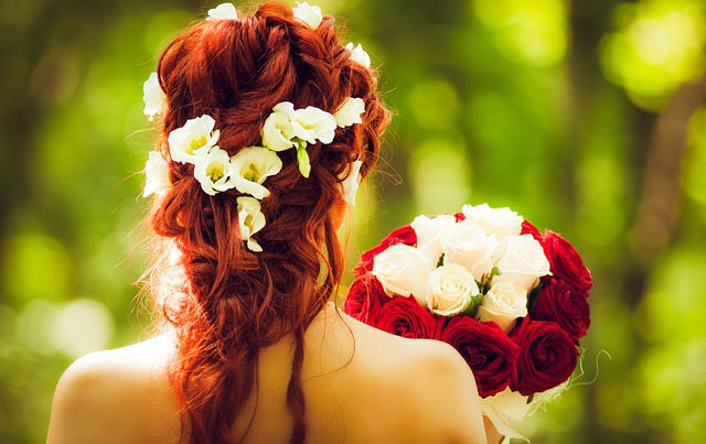 A red-haired bride facing a woody forest, while holding a red and white bouqet of roses.