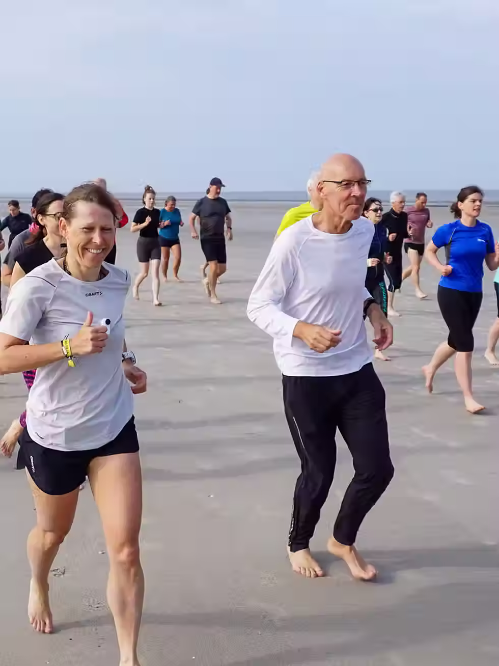 Eine Gruppe von Menschen, darunter eine Frau, rennt fröhlich am Strand entlang, im Hintergrund sind Wellen zu hören.