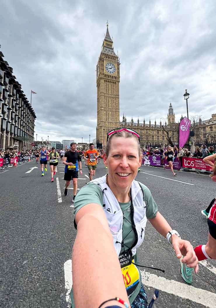 Läuferin macht Selfie beim Marathon in London vor Big Ben. Viele Teilnehmer im Hintergrund, grauer Himmel, bunte Kleidung, fröhliche Stimmung.