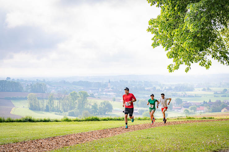 Drei Läufer auf einem Waldweg im Grünen, tragen bunte Shirts. Im Hintergrund hügelige Landschaft, bewölkter Himmel. Energetische Stimmung.