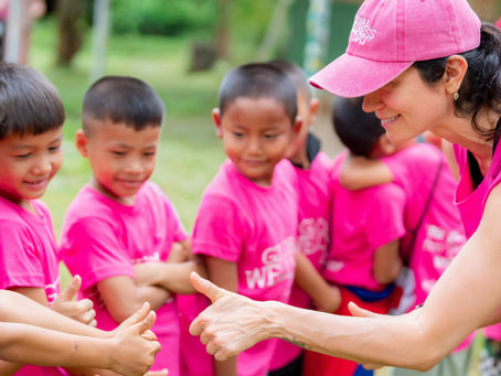 Woman in pink cap with Global Wellness Day logo gives thumbs-up to children in bright pink shirts outdoors.