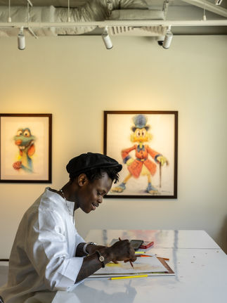 Artist sketching, inspired by displayed artwork of a frog and a gentleman in a red coat.