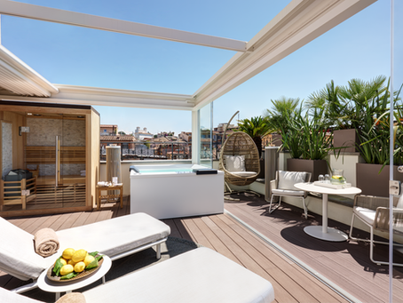 Rooftop terrace with sauna, hot tub, and loungers. Wicker chair, table with drinks, and greenery under a clear blue sky. Relaxing vibe.