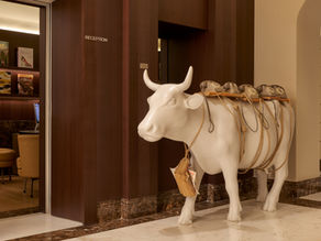 White cow sculpture with rotary phones on its back stands in a hotel reception area. Books and wooden decor are visible in the background.